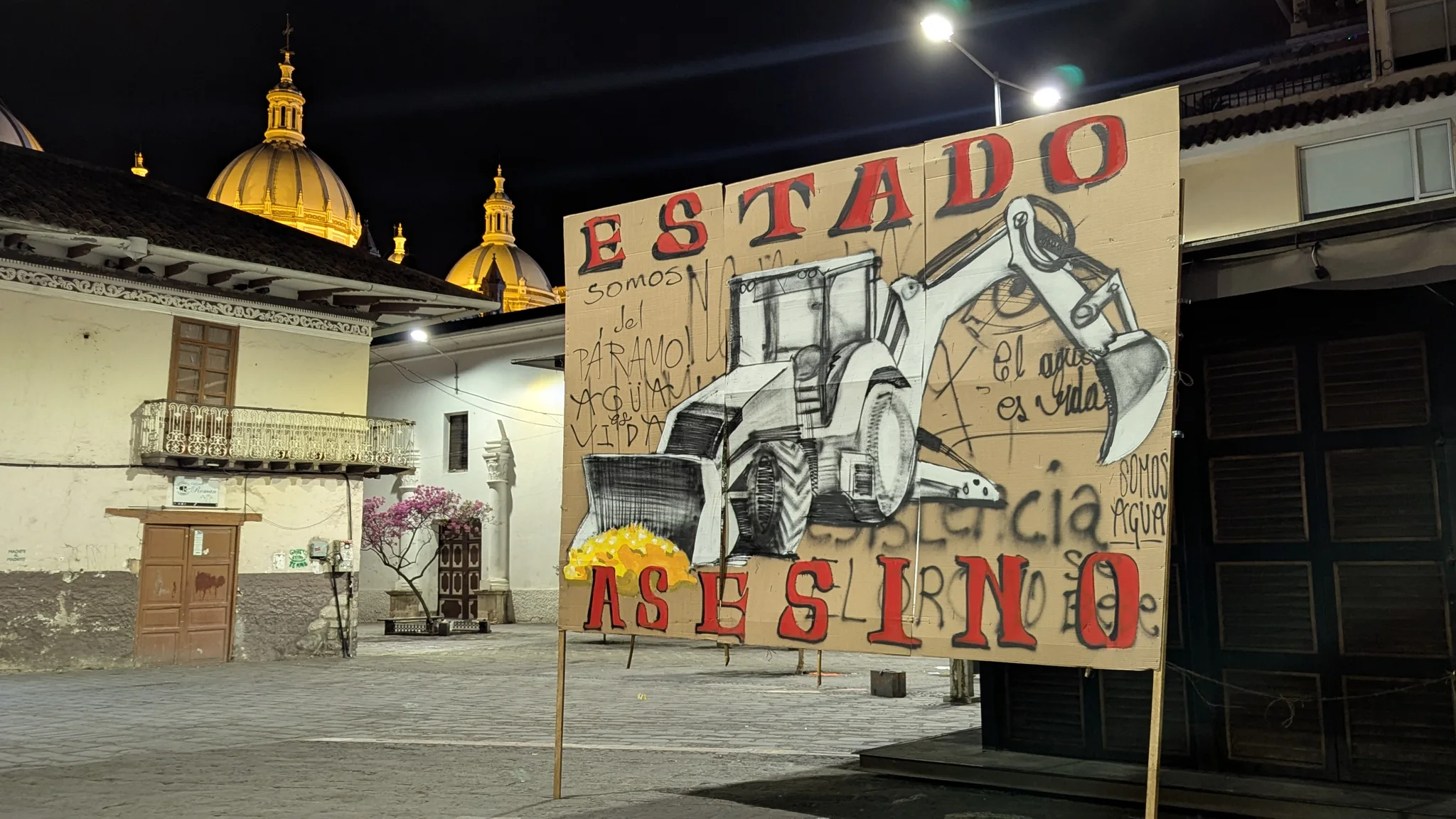 Un mural nocturno colorido y una torre de iglesia iluminan una plaza urbana.