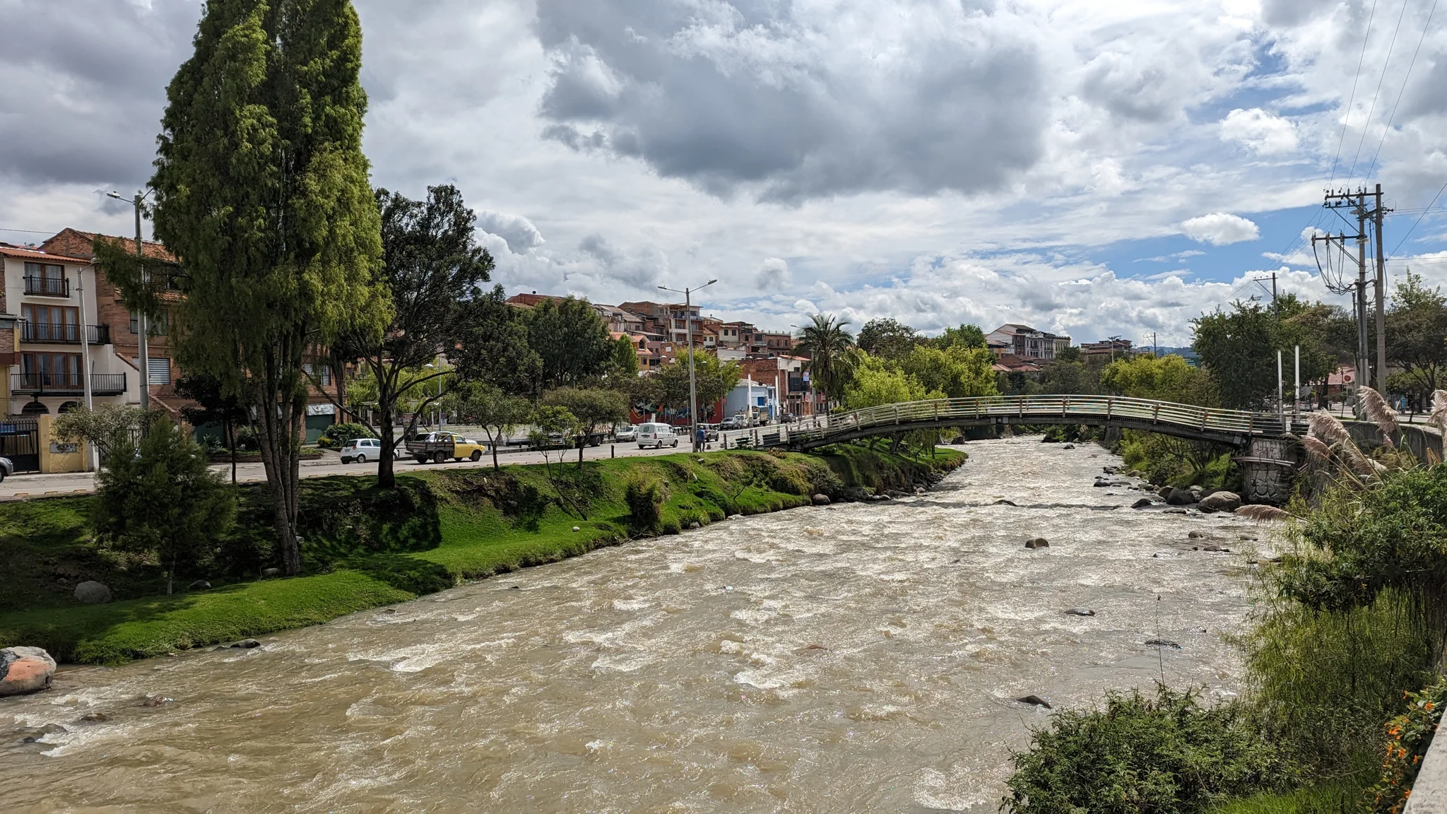 Un río de agua turbia cruza un parque urbano con árboles y edificios al fondo.