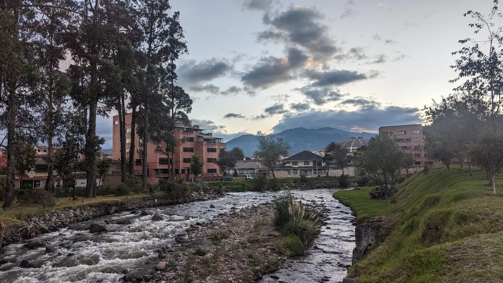 Una curva del río atraviesa un parque urbano con árboles y montañas al fondo.