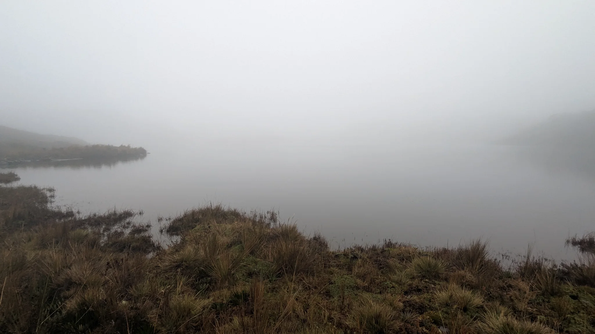 Una península oscura se pierde en la niebla sobre aguas tranquilas durante la salida de Osohuayco. El nombre de la laguna sigue sin confirmarse.