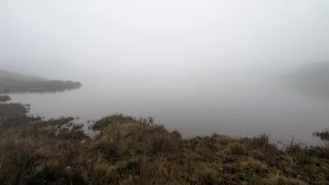 Una península oscura se pierde en la niebla sobre aguas tranquilas durante la salida de Osohuayco. El nombre de la laguna sigue sin confirmarse.
