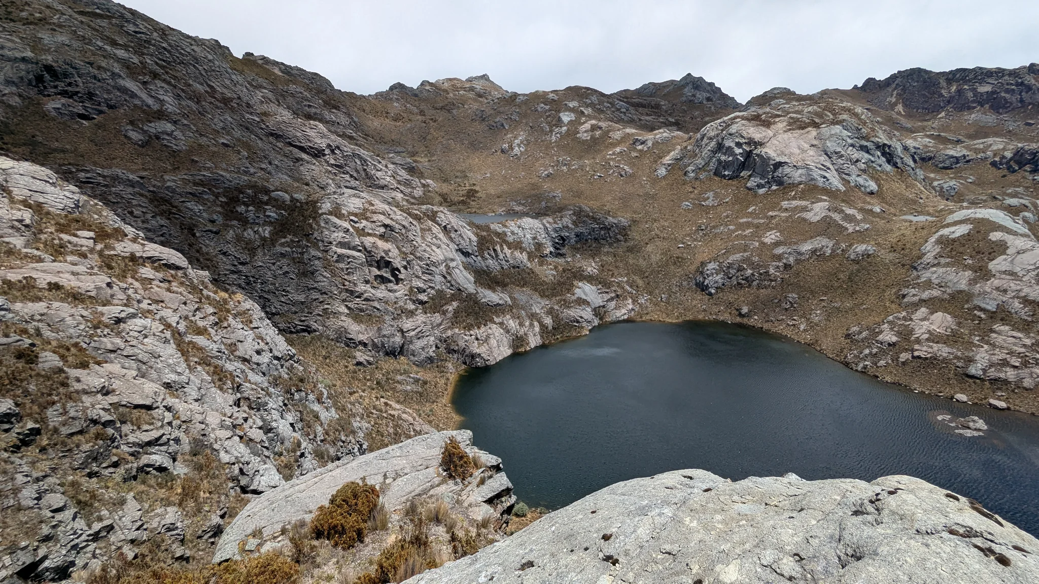 Una laguna oscura sin nombre confirmado ocupa una hondonada rocosa y escarpada bajo un amplio mirador de altura.