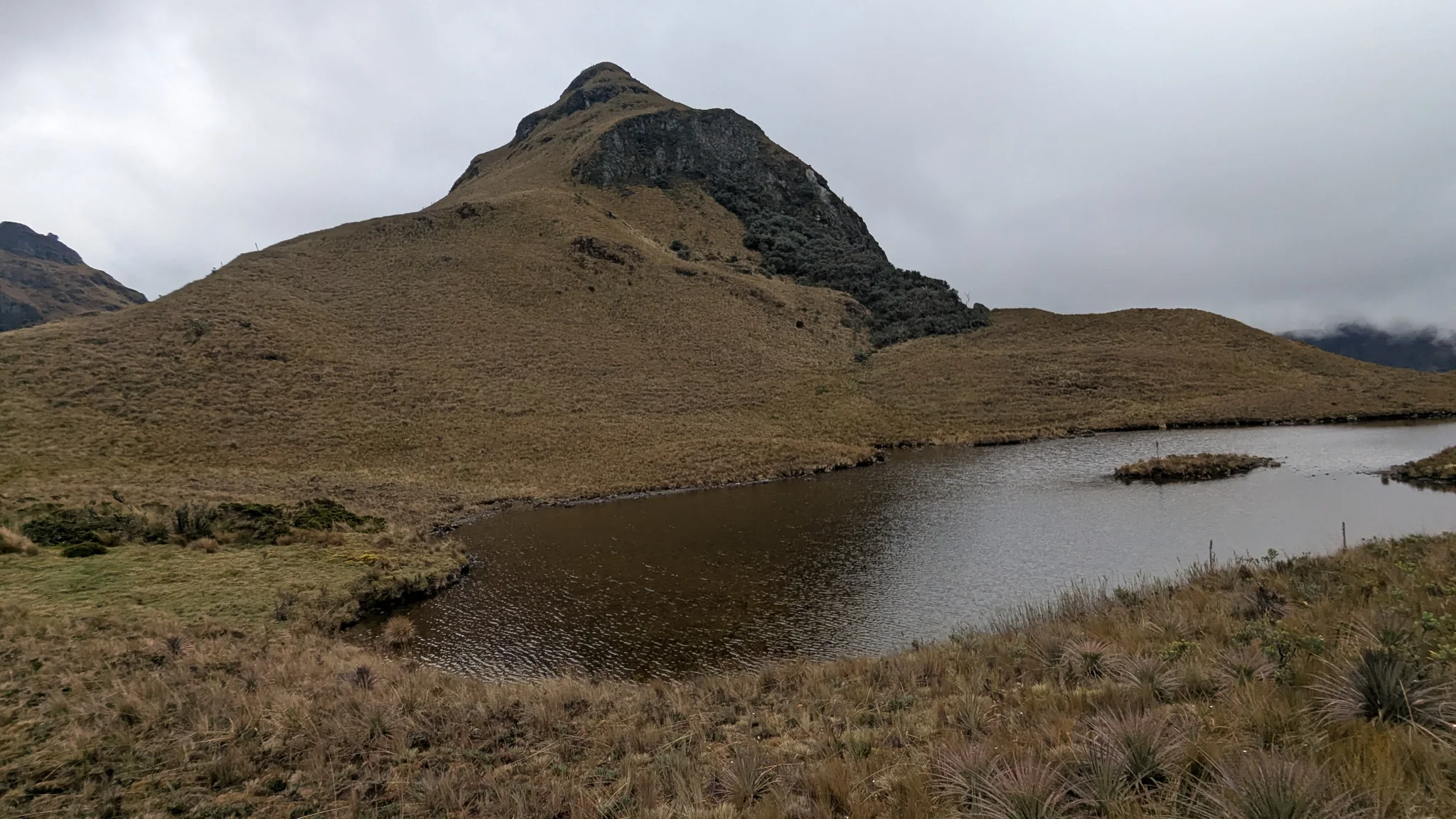 Una laguna oscura sin nombre confirmado se curva al pie de un cerro puntiagudo durante la travesía del Cajas.