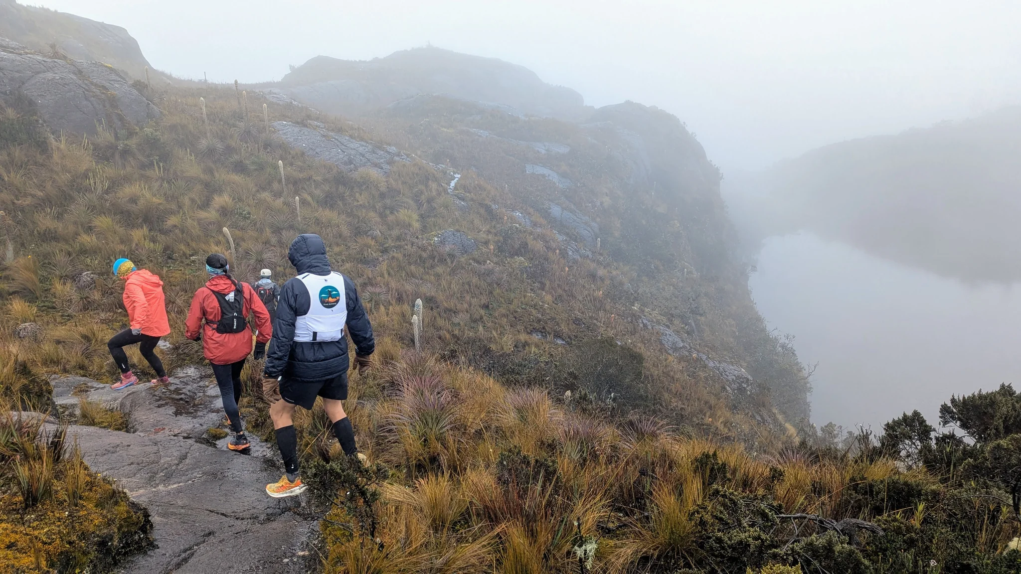 Unos caminantes descienden por un sendero rocoso junto al agua mientras la niebla oculta la orilla opuesta.