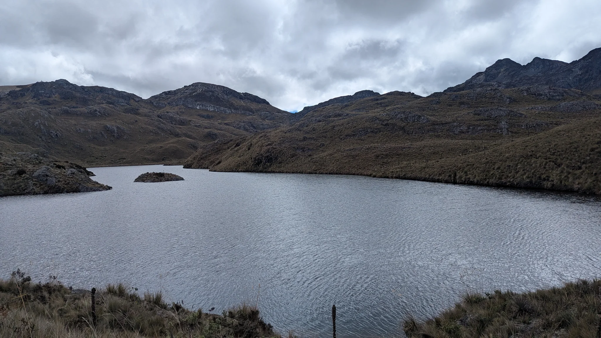 Un islote rocoso bajo sobresale de la laguna Riñoncocha, en una cuenca abierta cubierta de nubes.