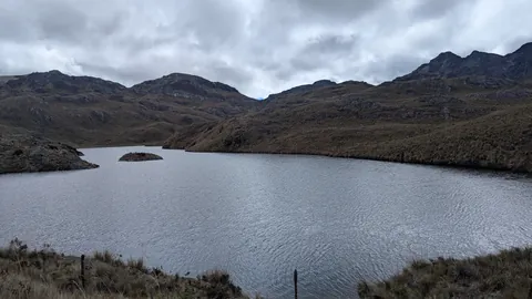Un islote rocoso bajo sobresale de la laguna Riñoncocha, en una cuenca abierta cubierta de nubes.