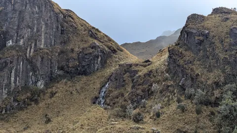 Una cascada blanca y fina desciende por una hendidura entre paredes de roca oscura y laderas de pajonal.
